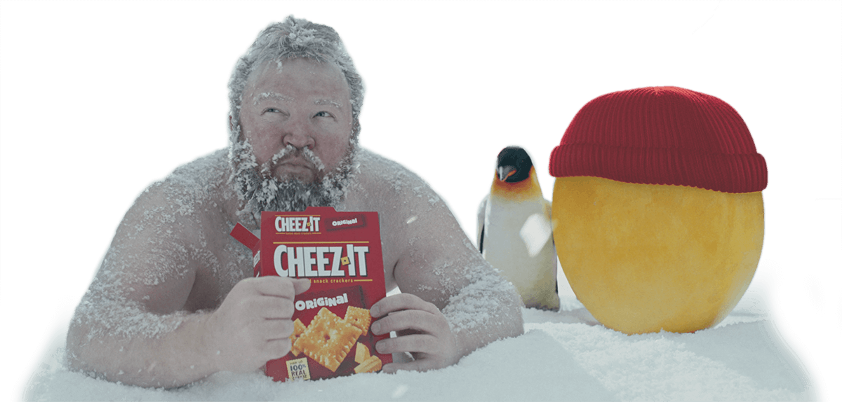 A shirtless man covered in snow eating Cheez-it crackers with a penguin and a cheeze wheel.
